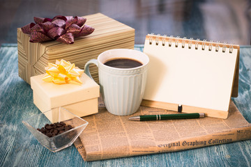 Festive still life with a cup of freshly brewed coffee, two gifts in white and light boxes, a vase with coffee beans, a notebook with a pen, a newspaper on a turquoise wooden table, a country