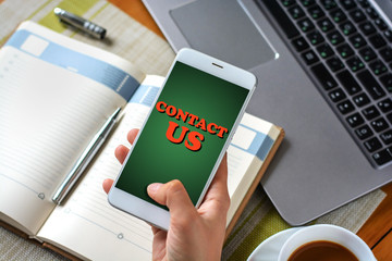 A woman's hand holds a smartphone with an inscription Contact us over a notebook, a cup of coffee and a laptop
