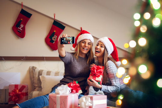 Two adorable excited girlfriends with Santa hat sitting on the bed for Christmas holidays one next to another and taking a selfie while laughing on laud.