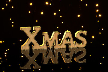 Wooden letters XMAS on a black reflective background. Behind the glowing garlands. Merry Christmas....