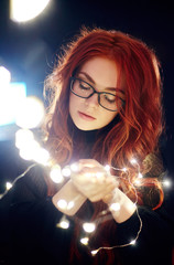 Art portrait of a woman with red hair in Christmas lights. Girl in glasses with reflected Christmas lights. Red hair in a yellow lights, tender feelings. Christmas came