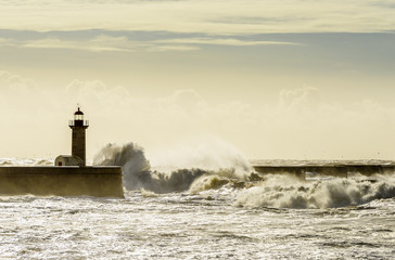 Landscape seascape lighthouse battered by huge waves on Atlantic Ocean with blue green skies and puffy clouds.
