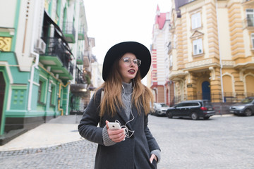 Stylish tourist girl strolling through the streets of the old city with headphones and smiles. Portrait of a happy girl with a phone in her hands on the streets of the city.