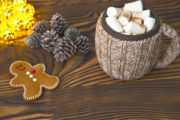 Cup of drink with marshmallow and tasty gingerbread cookie on Christmas lights and pines background