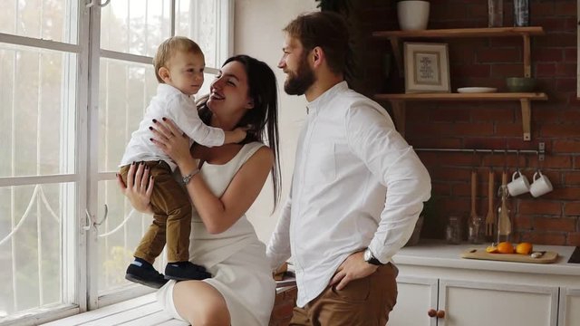 Young mother is holding her cute son sitting on the window sill together with young father. The window is decorated with Christmas wreath. Loving family together at home