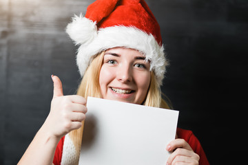 Portrait of a beautiful cheerful girl in Santa Claus hat red shirt and knitted scarf holding a white sheet of paper with place for text