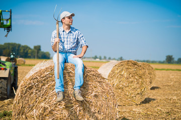 Naklejka na ściany i meble Smiling farmer sitting on an hay bale in his field