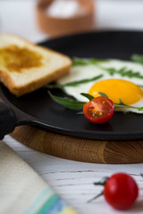 Chicken fried egg in a black frying pan with cherry tomatoes, arugula, salt. Breakfast on a white wooden table. Just cooked food