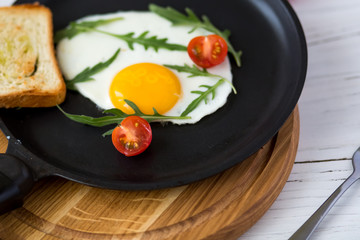 Chicken fried egg in a black frying pan with cherry tomatoes, arugula, salt. Breakfast on a white wooden table. Just cooked food