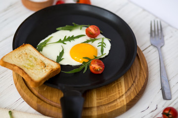 Chicken fried egg in a black frying pan with cherry tomatoes, arugula, salt. Breakfast on a white wooden table. Just cooked food