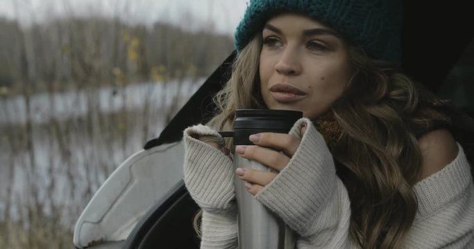 closeup portrait of handsome young woman / girl sitting outdoors in the car and warming up with hot beverage (cacao, coffee, tea) from thermos. she is wearing knitted cap, white sweater