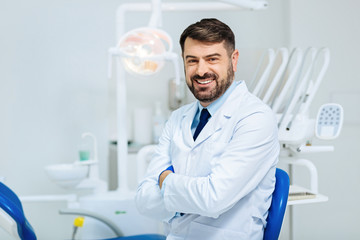 Healthy smile. Positive bearded dentist expressing cheer while standing in front of you and keeping arms in crossed position