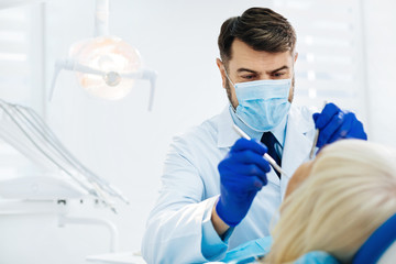 Professional of his business. Close up of experienced stomatologist with a medical face mask curing his patient while using dental tools