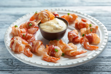 Plate with bacon-wrapped shrimps and tasty sauce on wooden table