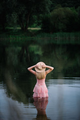 Beautiful blonde woman in a long pink dress and a hat standing in the summer river, enjoying the water, eyes closed