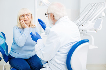Dentist consultation. Displeased woman sitting on the dental chair and touching her cheek while having a talk with stomatologist