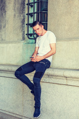 Obraz premium Eastern European College Student traveling, studying in New York. Wearing white T shirt, black jeans, sneakers, guy sitting on vintage wall on street, texting on cell phone. Instagram filtered effect.