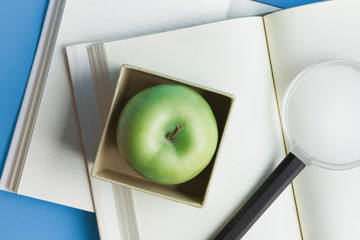Green apple in a box and magnifying glass on the open book, top view