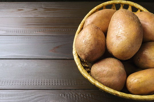 Uncooked, fresh crop of potatoes in a wicker basket on wooden table of dark brown planks background