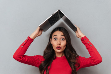 Shocked brunette woman in red blouse holding laptop computer overhead