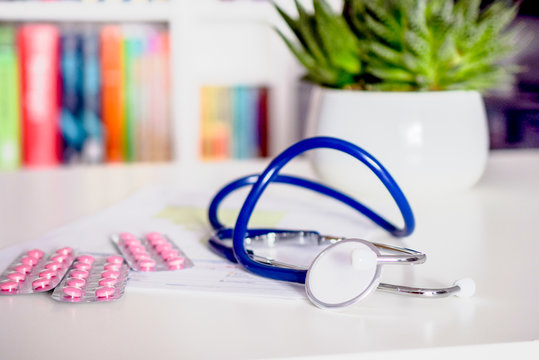 blue stethoscope and pink pills on white table in doctor's office or in hospital with chair and bookshelf in the background