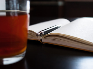 A glass mug with tea and a lemon slice, notebook with blank pages, black pen on dark desk