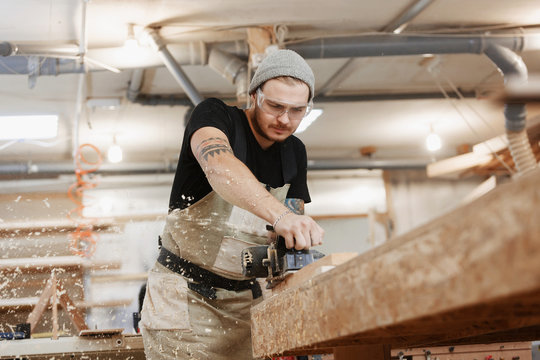 Carpenter working with electric planer on wooden plank in workshop. Hands and planer close up.