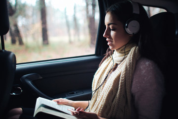 Woman in the car, autumn concept. Smiling pretty girl listening to music with headphones and reading a book moving in car