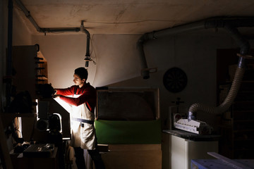 Fototapeta na wymiar Portrait of working young male carpenter wearing protective clothes with a beard and a red sweater handles the wood on a lathe in the workshop. dark lighting, carpenters workshop