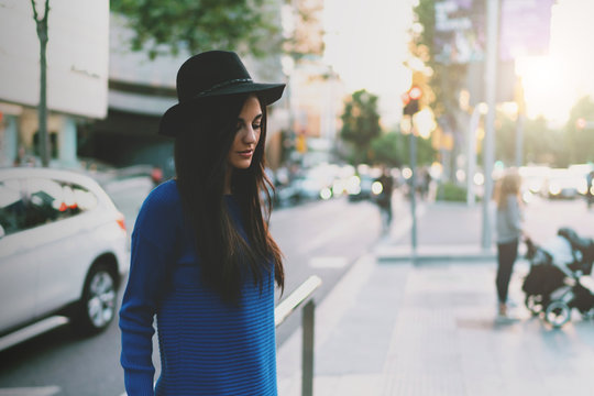 Portrait of a model look woman with long dark hair wearing hat and blue dress is looking down while standing on a vibrant city street. Stylish caucasian female standing on a blurred background.