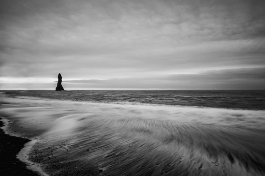 Coast of the Atlantic Ocean. Vic Beach in Iceland with black sand and a lonely rock in the sea. The wave ran up to the shore, a long exposure. Black and white photo.
