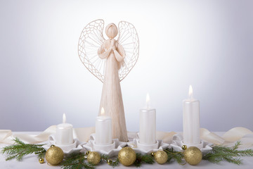 A white angel and four white Advent candles, Christmas tree branches are decorated with golden balls. The imminent background of Advent and Christmas