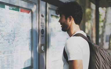 Naklejka premium Young bearded interantional student searching the route on a public transportation system map in unknown city. Handsome traveler with backpack looking at train timetable on a railway station.