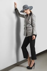 Pretty fashion model in jacket, pants and broad-brim hat with flowers posing near gray wall