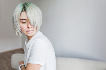 Portrait of cute  girl with white short  hairstyle  sitting on sofa in studio. She wears white dress. Her hair cover half  face. She is looking down.