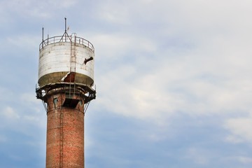 Water tower made of bricks, aluminum sheets, with metal pipes, glass small windows and a metal staircase against a cloudy white-blue sky.