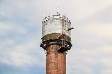 Water tower made of bricks, aluminum sheets, with metal pipes, glass small windows and a metal staircase against a cloudy white-blue sky.
