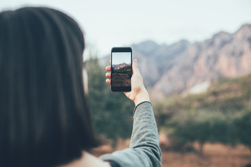 Young woman or girl makes photograph of mountains landscape on her smartphone camera, to share on internet social media platform through photography application for mobile device
