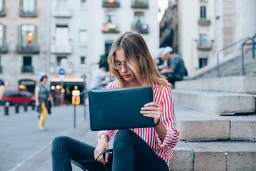 Young beautiful woman with blonde hair takes laptop in protective plastic mockup cover out of fashion leather bag. student sits on stairs or steps of university building, hipster freelance lifestyle