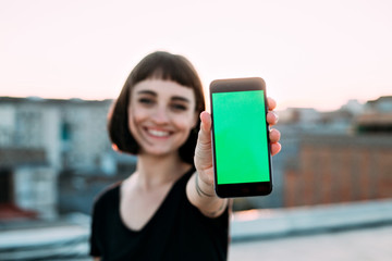 Happy smiling positive and cute young woman with unconventional natural beauty holds up to camera, smartphone with green chroma key screen for mockup, sunset light outdoors, concept apps