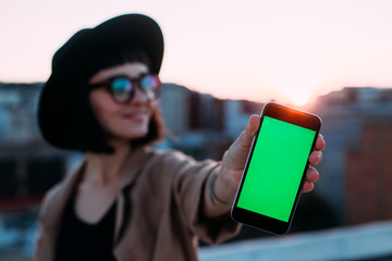 Beautiful young woman in fedora hat and hipster sunglasses, shows smartphone screen chroma key green background for mockup, smiles laughs, concept millennial gadget user and hipster internet addiction