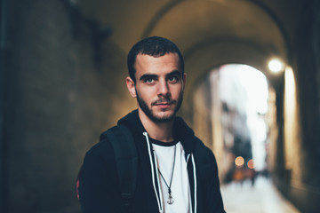 Obraz premium Handsome attractive young man in 20s, student or professional freelancer, looks into camera, stands in dark street or alley in Barcelona, exchange education program, confident and calm