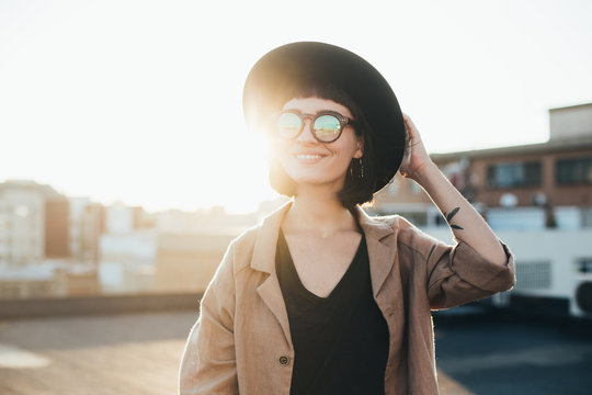 Cool and modern young beautiful teenager or woman poses to camera in sunset light on rooftop of building, wears reflective glasses with lenses, smiles and laughs happy and satisfied with lifestyle