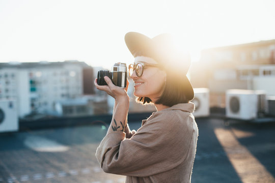 Cute adorable and trendy dressed in hipster outfit millennial young girl or woman makes photograph of sunset on vintage retro analog film camera, concept hobby and free time, happiness and chill