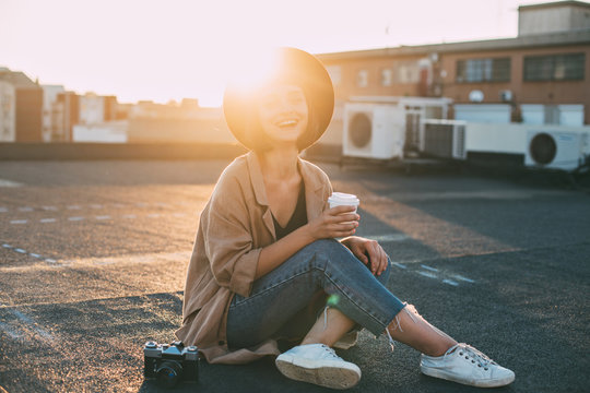 Happy trendy hipster woman in millennial cute outfit sits on ground at rooftop terrace, holds cup of take away coffee and looks into camera with smile, sunset light in her hair, concept forever young