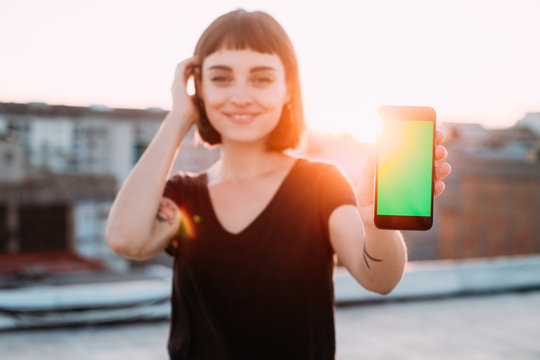 Happy smiling positive and cute young woman with unconventional natural beauty holds up to camera, smartphone with green chroma key screen for mockup, sunset outdoors, concept application development