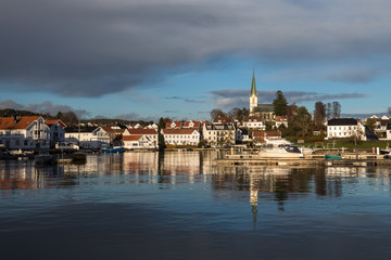 Naklejka na ściany i meble Lillesand, Norway - November 10, 2017: Lillesand City seen from the harbor. Blue sky and clouds, reflections of city in the ocean.