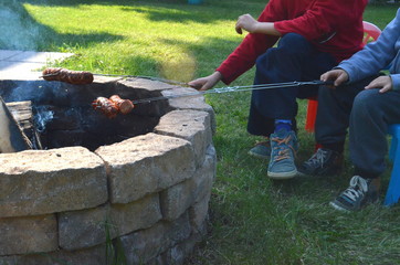 Two boys are grilling or barbecuing Polish sausages over an open fire pit in their backyard