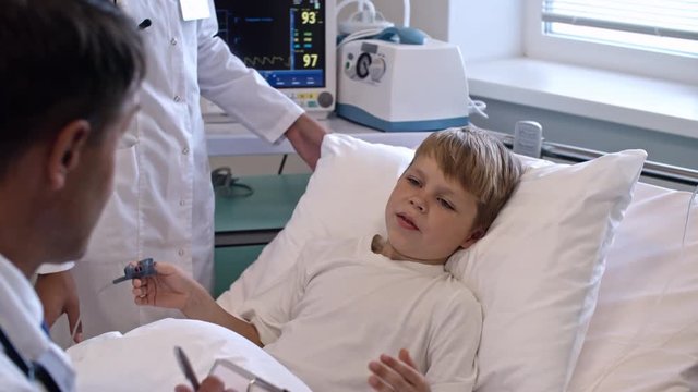 Zoom out of little boy lying on hospital bed and speaking with male doctor while nurse tracking his pulse with medical monitor