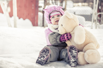 Beautiful baby embracing toy plush bear sitting on snow in park in cold sunny winter day  during winter holidays
